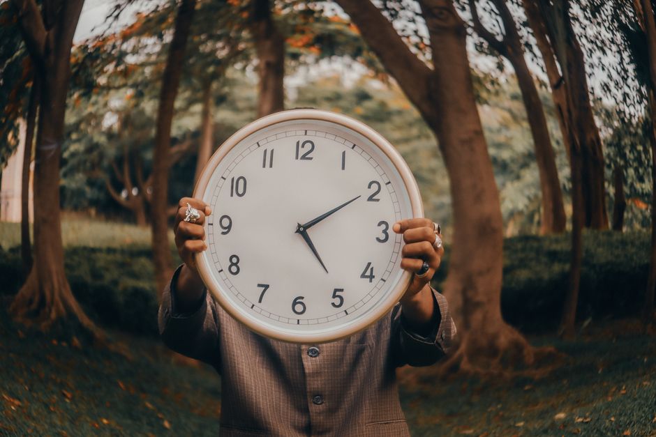 An individual holding a large clock in an outdoor forest environment, symbolizing time.